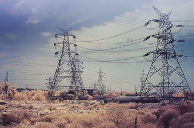 Low angle view of electricity pylons on field against cloudy sky during sunny day