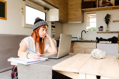 Focused female traveler sitting at table in truck and taking notes while working on project remotely and leaning on hand