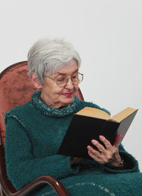 Senior woman reading book while sitting on chair against white background