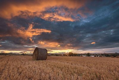 Hay bales on agricultural field against cloudy sky during sunset