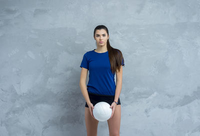 Portrait of young woman holding volleyball standing against wall