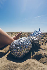 Low section of woman legs by pineapples at beach against clear blue sky