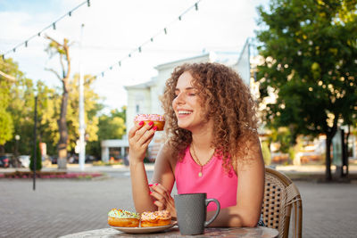 Young attractive curly woman sits at table in cafe on summer terrace. colorful delicious donuts, 