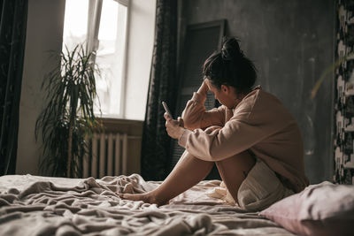 Midsection of woman holding mobile phone while sitting on bed at home