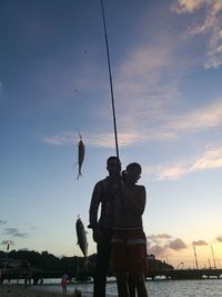 Silhouette people holding fish in rod while standing against sky during sunset