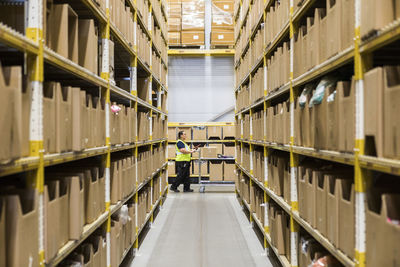 Full length side view of mature female warehouse worker pushing cart seen through narrow aisle in industrial building