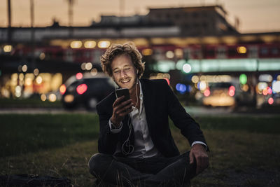 Smiling businessman sitting on meadow at dusk with cell phone and earphones