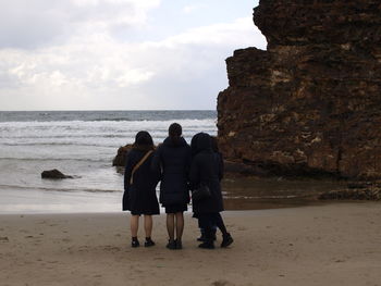 Rear view of friends standing on shore at beach against sky