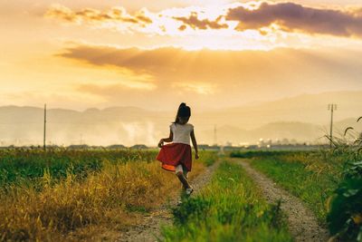 Girl standing on country road amidst farms against cloudy sky during sunset