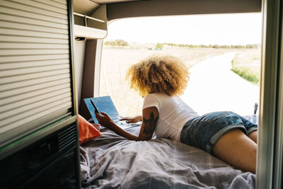 Young african american female traveler with curly hair working on laptop while lying down inside camper van during summer holidays