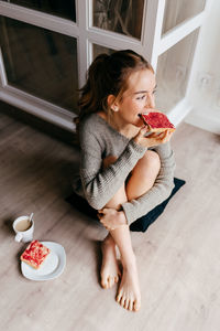 From above of positive young female in casual wear having breakfast with sweet toast and cup of coffee while sitting on floor in room