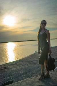 Full length portrait of young woman standing at beach during sunset