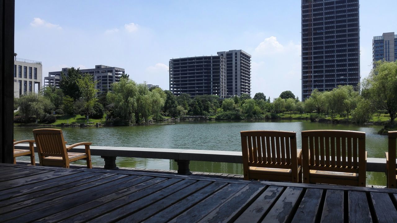 water, building exterior, tree, built structure, architecture, sky, city, railing, lake, bench, chair, relaxation, river, day, tranquility, sunlight, skyscraper, nature, outdoors, pier