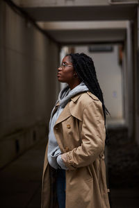 Side view of young beautiful african american female with braids in casual outfit and glasses looking away while standing in hallway of contemporary building