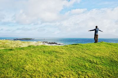 Rear view of man with arms outstretched standing on grass field against sky