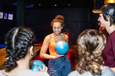 Smiling teenage girl holding bowling ball while standing amidst friends