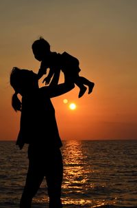 Silhouette mother picking up child while standing by sea against sky during sunset