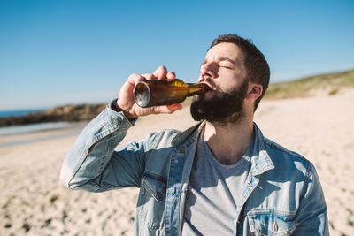 Young man drinking alcoholic drink bottle while standing at beach during sunny day