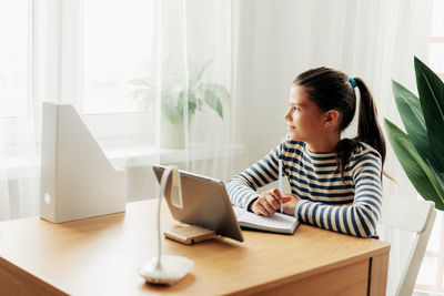 Happy pensive schoolgirl sits at the table at home, does homework on the tablet 