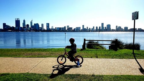 Side view of boy riding bicycle on footpath by river against buildings in city