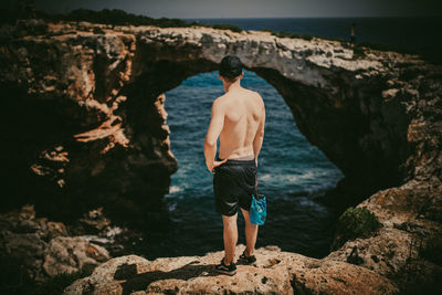 Rear view of shirtless man standing on rock by sea