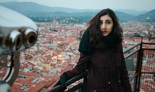 Portrait of beautiful woman standing by railing against cityscape