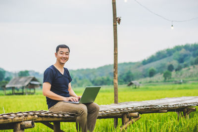 Portrait of man using laptop while sitting on boardwalk at rice paddy