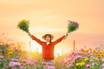 Portrait of woman standing by flowering plants against sky during sunset