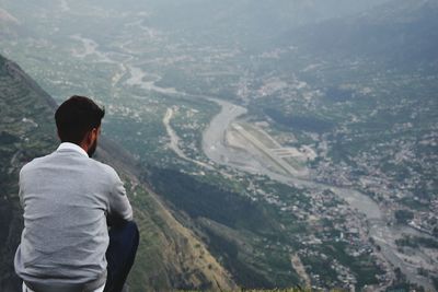 Rear view of man sitting on cliff looking at townscape