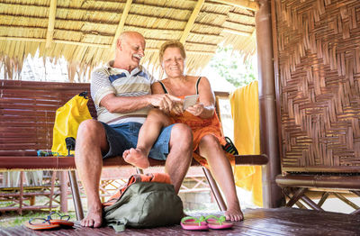 Smiling senior couple using phone while sitting on bench in hut