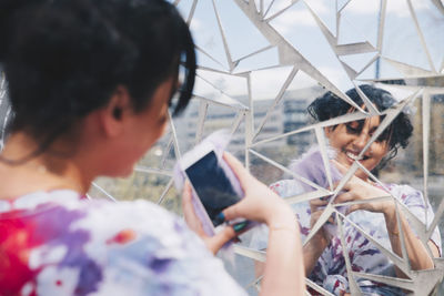 Young woman taking selfie through mobile phone against glass mosaic wall