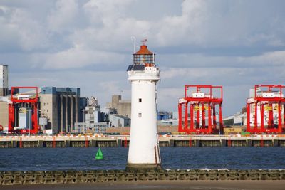 Lighthouse amidst buildings by sea against sky