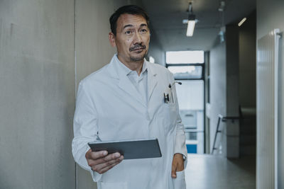 Scientist with hand in pocket using digital tablet while standing at clinic corridor