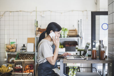 Female entrepreneur talking over smart phone while working on laptop at store