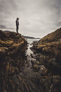 Man standing on cliff at beach against cloudy sky