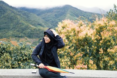 Smiling woman with umbrella sitting on retaining wall against mountains