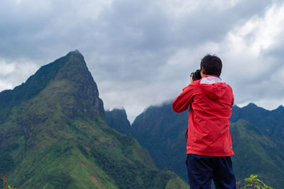 Rear view of man photographing mountains against cloudy sky
