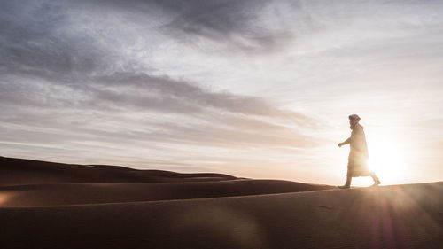 Man walking at desert against sky during sunset