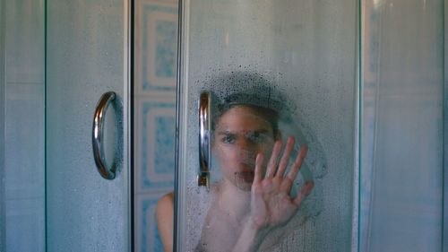 Portrait of shirtless young man looking through condensed window