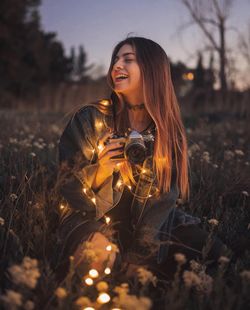 Young woman smiling while holding camera and illuminated string lights while sitting amidst plants on land 