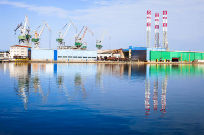 Reflection of cranes at commercial dock in sea against sky