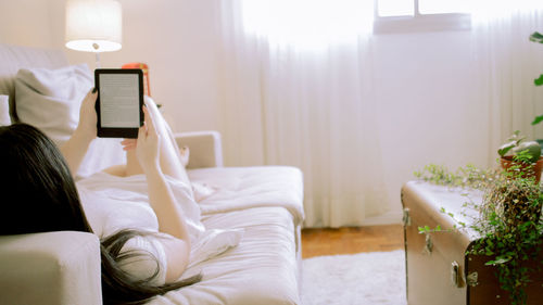  woman laying down on the couch, reading on her e-reader, with natural light in the living room