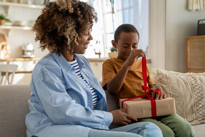 Pleased african american child boy happy to open gift from caring mom unties ribbon in anticipation