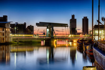 Illuminated city by boats moored on river against sky at night