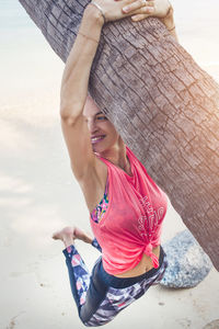 Smiling mid adult woman hanging on palm tree at beach