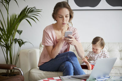 Fashion designer using mobile phone while sitting with daughter on sofa at home