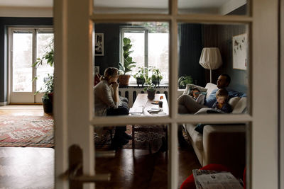 Family using various technologies in living room seen through transparent door at home