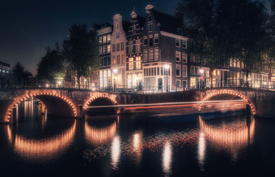 Illuminated bridge over river by buildings in city at night
