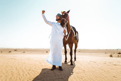 Man raising hand while standing with horse in dessert against sky