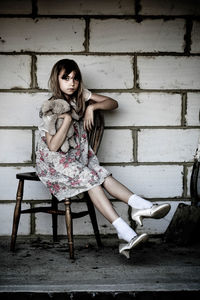 Portrait of girl holding stuffed toy and sitting on chair against wall